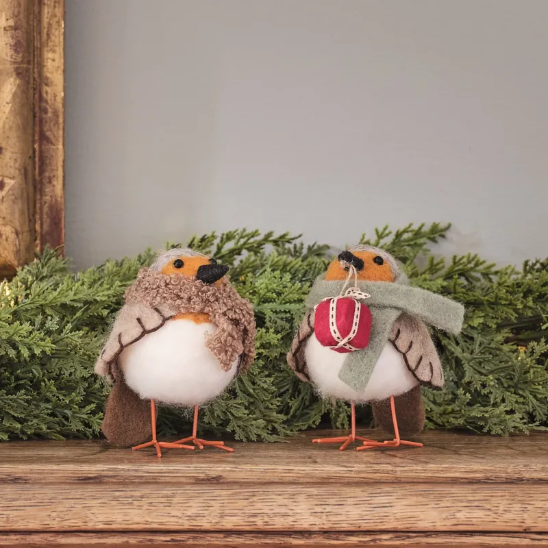 Zwei cremefarbene Vogel-Dekofiguren mit roten gestrickten Weihnachtsmützen und burgundy Schleife, stehend auf grünem Moosuntergrund mit Holzstamm-Hintergrund – niedliche Weihnachtsfiguren für Tischdekoration, Kaminsims und festliche Wohnzimmer-Arrangements mit warmem Charme.