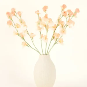 Die cremefarbene Keramikvase enthält den Kunstblumenstrauß Pea Peach aus Seide mit zarten pfirsichfarbenen und hellrosa Blüten mit langen grünen Stielen vor einem hellen, neutralen Hintergrund.