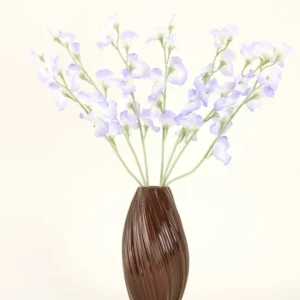 In einer braunen, gerippten Vase befindet sich der künstliche Seidenblumenstrauß Erbsen-Lavendel mit zarten Blütenblättern und grünen Stielen vor einem schlichten, cremefarbenen Hintergrund für eine dezente Landhausatmosphäre.