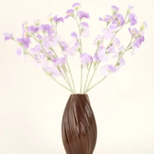 Eine strukturierte braune Vase hält den künstlichen Seidenblumenstrauß Erbsenlila-hellviolette Seidenblumen mit grünlichen Stielen vor einem schlichten, hellen Hintergrund.