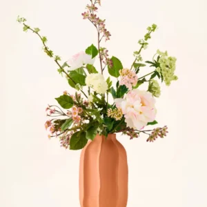 In einer pfirsichfarbenen Keramikvase befindet sich ein Strauß rosa-weißer Kunstblumen aus Seide, der „Dreamy Meadow“-Strauß, mit grünen Blättern und Stielen vor einem schlichten, hellen Hintergrund.