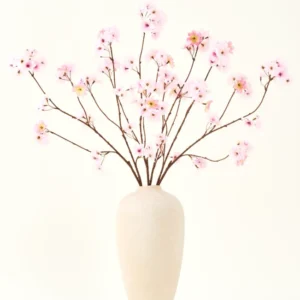 Der Seiden-Kunstblumenstrauß Kirschblütenrosa 114 cm hat lange, dünne Zweige mit Büscheln hellrosa Blüten in einer beigen Keramikvase, die einen auffälligen Kontrast zum schlichten weißen Hintergrund bilden.