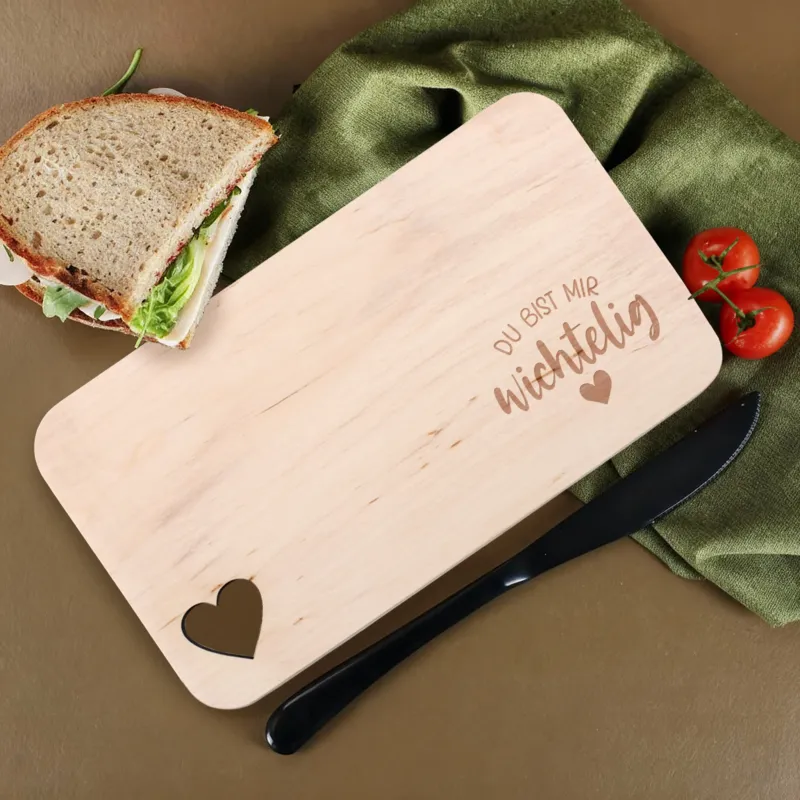 Holzbrettchen mit Gravur „Du bist mir wichtelig“ und Herzausschnitt, dekoriert mit Sandwich und Tomaten.