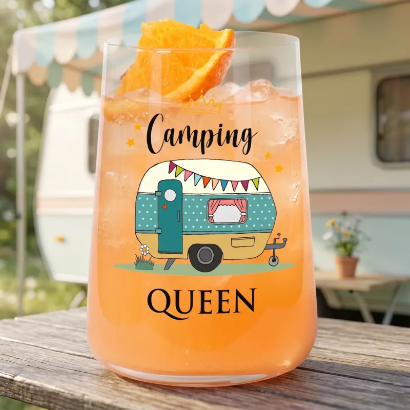 Trinkglas mit Camping Queen Wohnwagen-Motiv vor Wohnwagen-Hintergrund und orangefarbenem Getränk