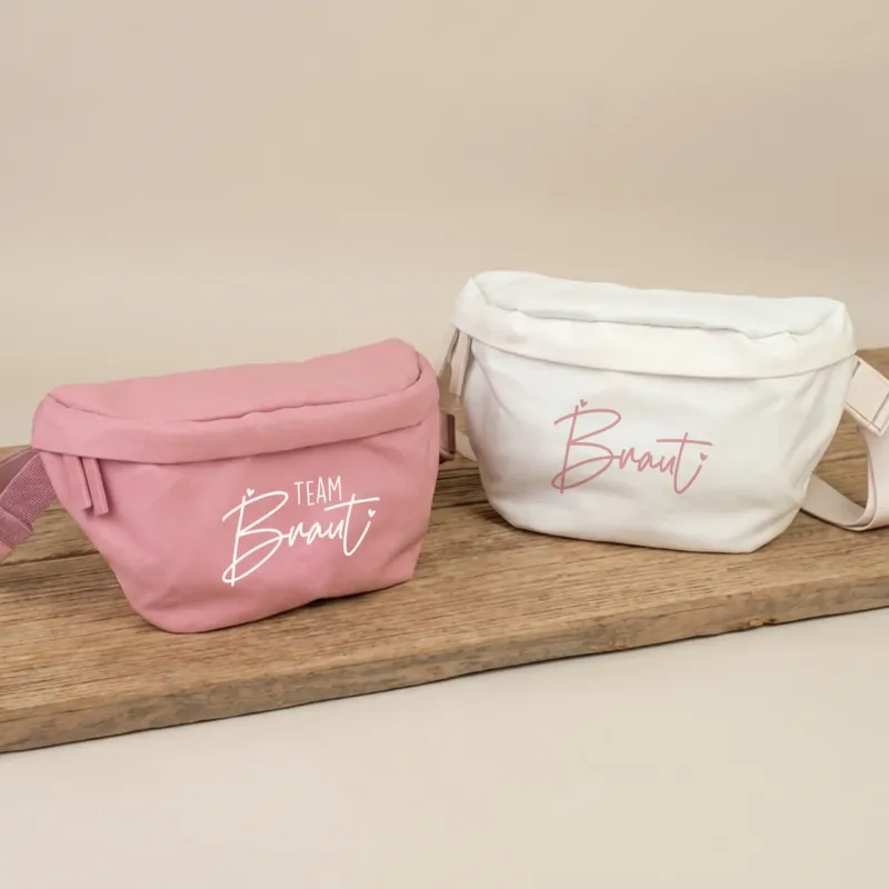 Rosa JGA Bauchtasche mit Aufdruck Team Braut und cremefarbene Bauchtasche mit Aufdruck Braut auf Holzbrett