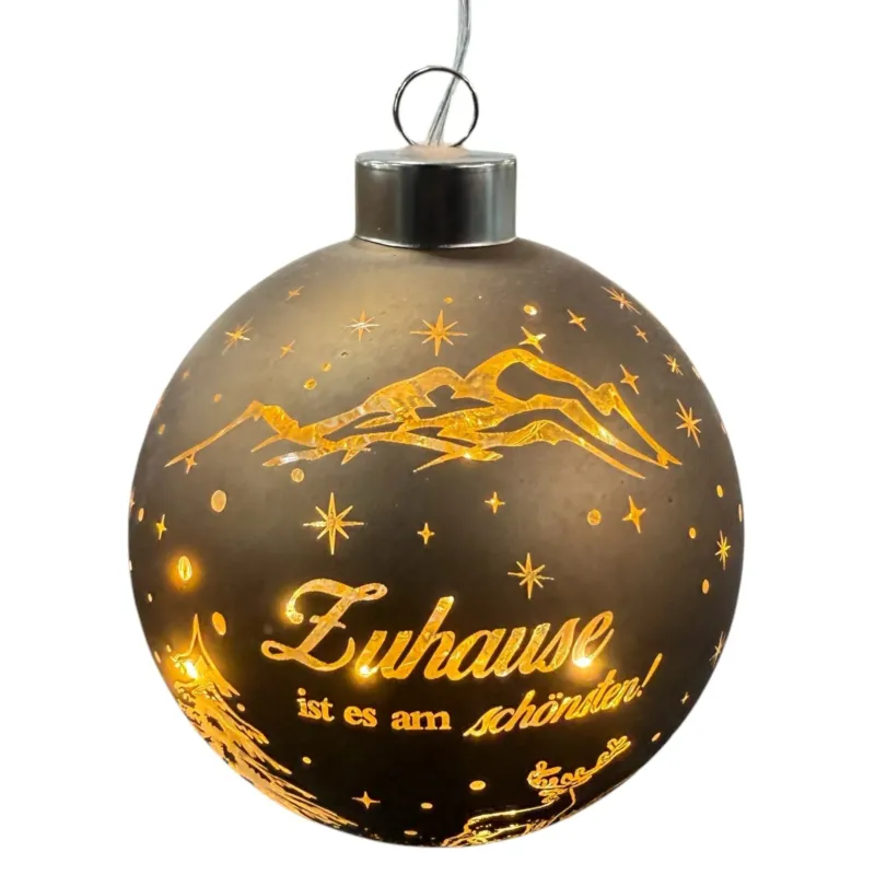 Eine runde, braune LED-Dekokugel zum Aufhängen mit warmweißem Licht zeigt eine winterliche Szene mit Bergen, Tannenbaum, Hirsch und dem Schriftzug „Zuhause ist es am schönsten!“.