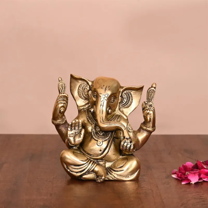 A brass figurine of Ganesha, the elephant-headed god, with big ears, seated in a gold-colored finish, displayed on a flat surface.
